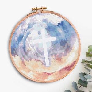 Pode incluir: Um bastidor de bordado em ponto cruz com uma cruz branca sobre um fundo de redemoinhos azuis, p&ecirc;ssego e brancos. O bastidor de madeira redondo tem um parafuso de ajuste de lat&atilde;o. A obra de arte evoca espiritualidade e f&eacute;.