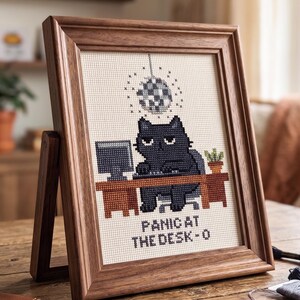 Puede incluir: Un bordado en punto de cruz enmarcado que muestra un gato negro sentado en un escritorio con una computadora y una bola de discoteca arriba. El texto PANIC AT THE DESK-O est&aacute; bordado en la parte inferior. La pieza tiene un marco de madera oscura.