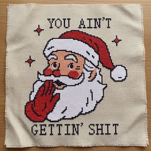 May include: A cross-stitch embroidery on beige Aida fabric featuring a Santa Claus head wearing a red hat with a white pom-pom. Santa has a white beard, red cheeks, and a red gloved hand covering his mouth. The text reads YOU AIN'T GETTIN' SHIT.