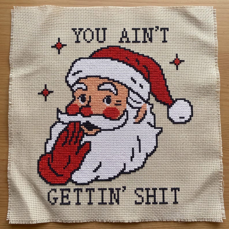 May include: A cross-stitch embroidery on beige Aida fabric featuring a Santa Claus head wearing a red hat with a white pom-pom. Santa has a white beard, red cheeks, and a red gloved hand covering his mouth. The text reads YOU AIN'T GETTIN' SHIT.