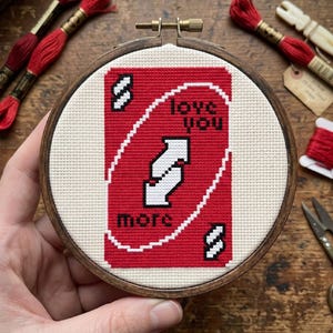 Pu&ograve; includere: Un telaio da ricamo circolare in legno contenente un disegno a punto croce di una carta da gioco rossa. La carta presenta un simbolo a doppia freccia bianca al centro con il testo nero "love you" sopra e "more" sotto, e la lettera "S" negli angoli.