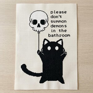 May include: A cross-stitch embroidery on off-white Aida fabric featuring a black cat holding a string attached to a white skull balloon. The text reads, please don't summon demons in the bathroom, stitched in black thread in a pixelated font style.