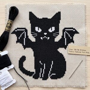 Puede incluir: Un patr&oacute;n de punto de cruz sobre tela Aida natural de 14ct que muestra un gato negro con alas de murci&eacute;lago, ojos blancos y colmillos. El texto dice: Size: 78x78 Stitches, Fabric: Natural 14ct Aida. Incluye hilo de bordar negro y una aguja.