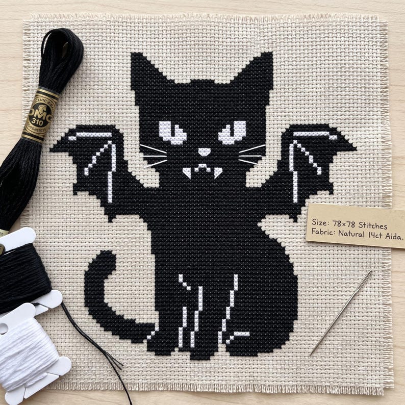 Puede incluir: Un patr&oacute;n de punto de cruz sobre tela Aida natural de 14ct que muestra un gato negro con alas de murci&eacute;lago, ojos blancos y colmillos. El texto dice: Size: 78x78 Stitches, Fabric: Natural 14ct Aida. Incluye hilo de bordar negro y una aguja.