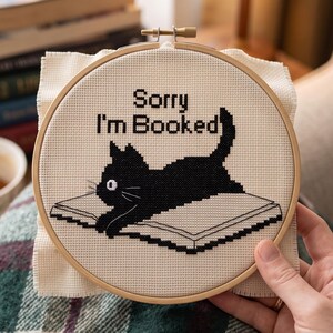 May include: A circular wooden embroidery hoop containing a cross-stitch design on cream-colored fabric. The design features a black cat lying on an open book with the black text "Sorry I'm Booked" stitched above the cat in a pixelated font style.