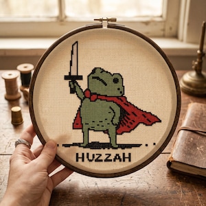 May include: A circular wooden embroidery hoop containing a cross-stitch pattern of a green frog wearing a red cape and holding a sword aloft. The text HUZZAH is stitched in black pixelated letters at the bottom of the cream-coloured fabric.