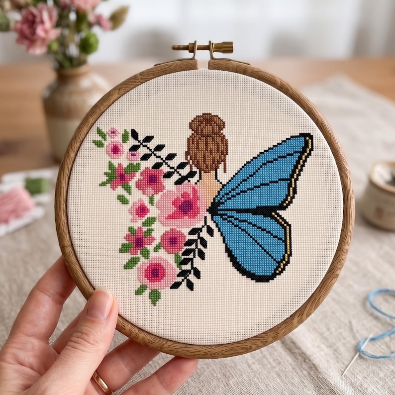 Peut inclure: Un projet de broderie au point de croix dans un tambour en bois rond repr&eacute;sentant une personne avec un chignon brun. Un c&ocirc;t&eacute; du motif montre des fleurs roses avec des feuilles vertes, tandis que l'autre affiche une grande aile de papillon bleue.