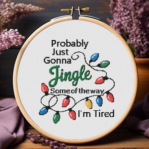 Może przedstawiać: Tamborek do haftu krzyżykowego z napisem "Probably Just Gonna Jingle Some of the way, I'm Tired." Projekt przedstawia kolorowe lampki choinkowe w kolorze czerwonym, niebieskim, zielonym i ż&oacute;łtym. Tamborek jest wykonany z drewna i ma mosiężną śrubę.
