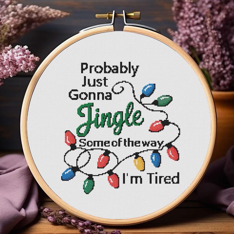 Może przedstawiać: Tamborek do haftu krzyżykowego z napisem "Probably Just Gonna Jingle Some of the way, I'm Tired." Projekt przedstawia kolorowe lampki choinkowe w kolorze czerwonym, niebieskim, zielonym i ż&oacute;łtym. Tamborek jest wykonany z drewna i ma mosiężną śrubę.