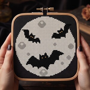 Pu&ograve; includere: Un telaio da ricamo quadrato in legno contenente un disegno a punto croce di una grande luna pallida e craterizzata su sfondo nero. Tre pipistrelli neri con piccoli occhi arancioni sono posizionati sulla luna, che presenta macchie grigio chiaro.