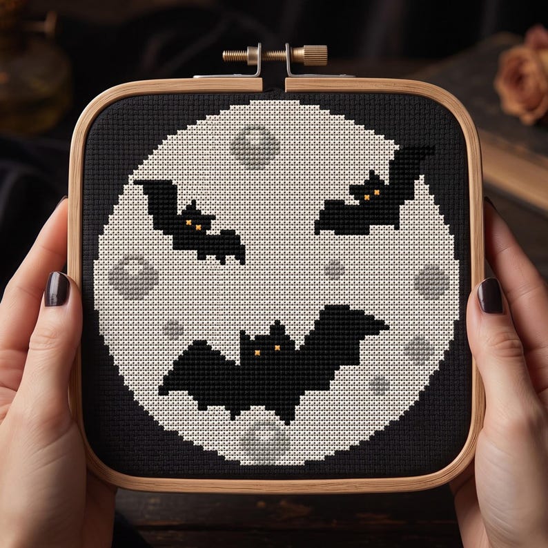 Pu&ograve; includere: Un telaio da ricamo quadrato in legno contenente un disegno a punto croce di una grande luna pallida e craterizzata su sfondo nero. Tre pipistrelli neri con piccoli occhi arancioni sono posizionati sulla luna, che presenta macchie grigio chiaro.