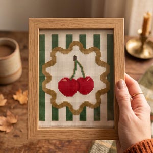 May include: A framed cross-stitch embroidery featuring two bright red cherries with green stems centred on a white scalloped background. The design is set against a backdrop of vertical dark green and white stripes, all encased in a light wood frame.