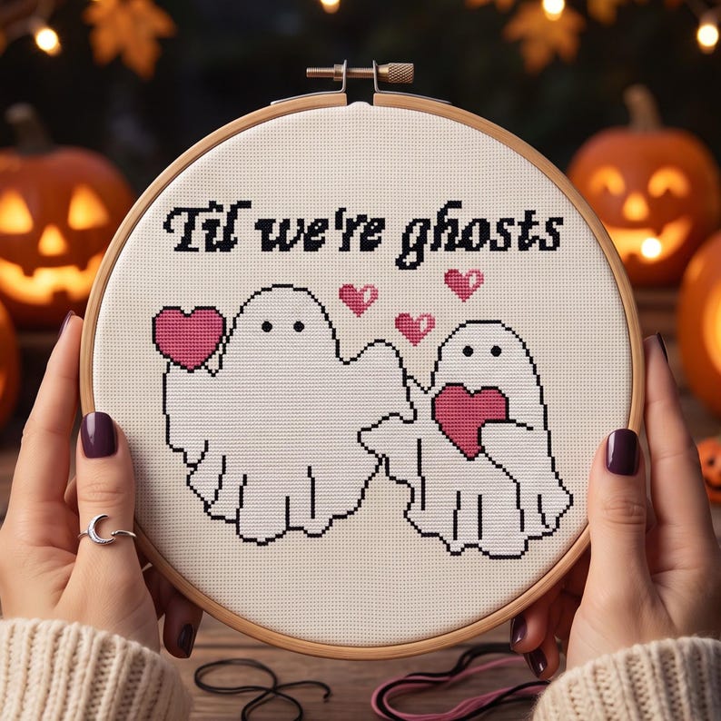 Pode incluir: Um bastidor de bordado redondo de madeira contendo tecido branco com um design em ponto cruz de dois fantasmas brancos segurando cora&ccedil;&otilde;es cor-de-rosa. O texto "Til we're ghosts" est&aacute; bordado a preto acima, com tr&ecirc;s pequenos cora&ccedil;&otilde;es a flutuar.