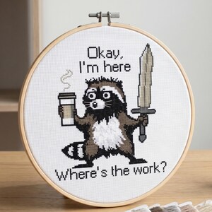 Puede incluir: Arte de punto de cruz en un aro que muestra un mapache con una taza de caf&eacute; y una espada. El texto dice: "Okay, I'm here. Where's the work?" El mapache es marr&oacute;n, negro y blanco.