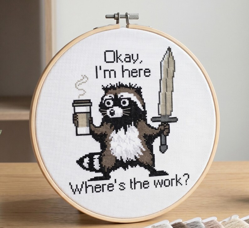 Puede incluir: Arte de punto de cruz en un aro que muestra un mapache con una taza de caf&eacute; y una espada. El texto dice: "Okay, I'm here. Where's the work?" El mapache es marr&oacute;n, negro y blanco.