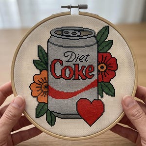 May include: A cross-stitch embroidery in a round wooden hoop featuring a silver soda can with the text Diet Coke in red and black lettering. The can is surrounded by orange and red flowers with green leaves and a solid red heart at the bottom.