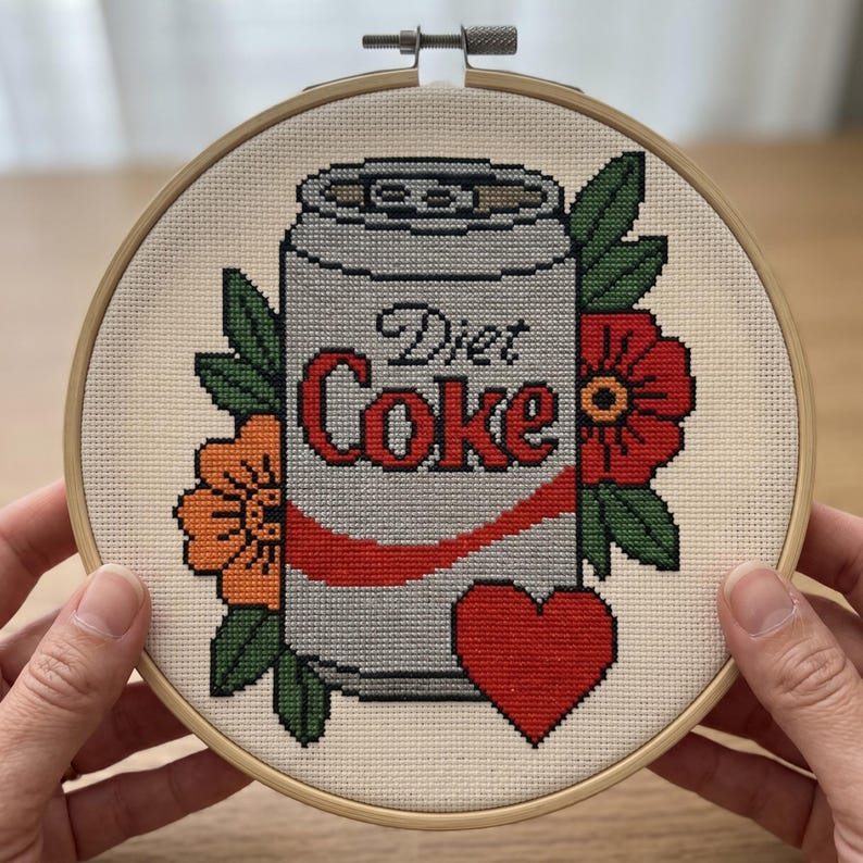 May include: A cross-stitch embroidery in a round wooden hoop featuring a silver soda can with the text Diet Coke in red and black lettering. The can is surrounded by orange and red flowers with green leaves and a solid red heart at the bottom.