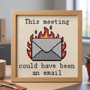 Op de afbeelding: Een vierkant ingelijst kruissteek borduurwerk met een grijze envelop omringd door oranje en gele vlammen. De zwarte geborduurde tekst boven en onder de envelop luidt, This meeting could have been an email, in een lichthouten lijst.