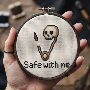 May include: A circular wooden embroidery hoop containing a cross-stitch design on cream-colored fabric. The design features a beige skull attached to a brown safety pin with a small lightning bolt icon and the black text "Safe with me".