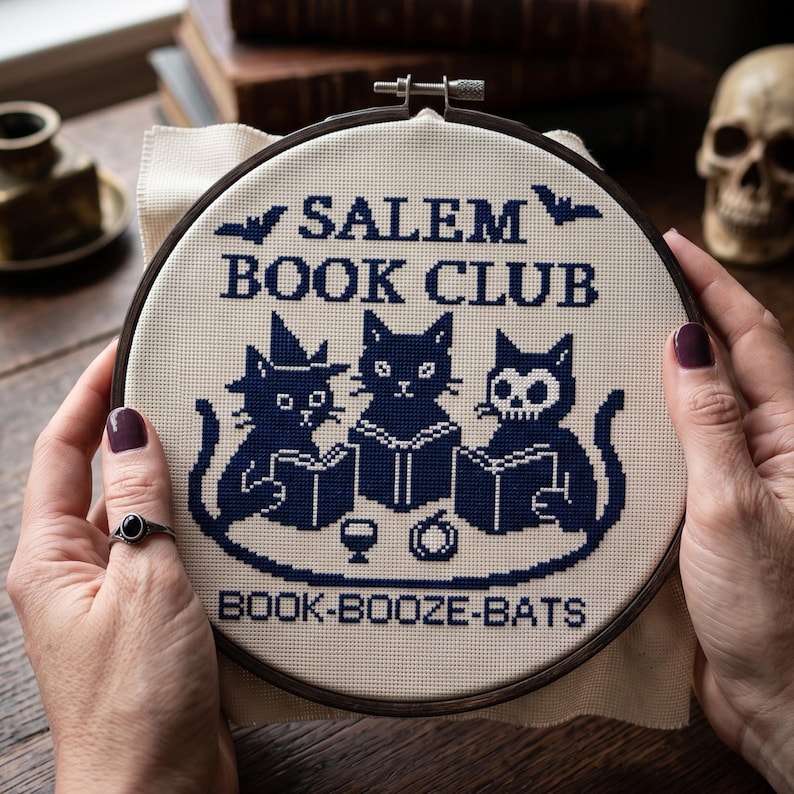 Puede incluir: Un bastidor de bordado circular con tela color crema y un dise&ntilde;o de punto de cruz azul oscuro. El texto dice SALEM BOOK CLUB y BOOK-BOOZE-BATS. La imagen muestra tres gatos leyendo libros, dos murci&eacute;lagos, una copa de vino y un anillo.