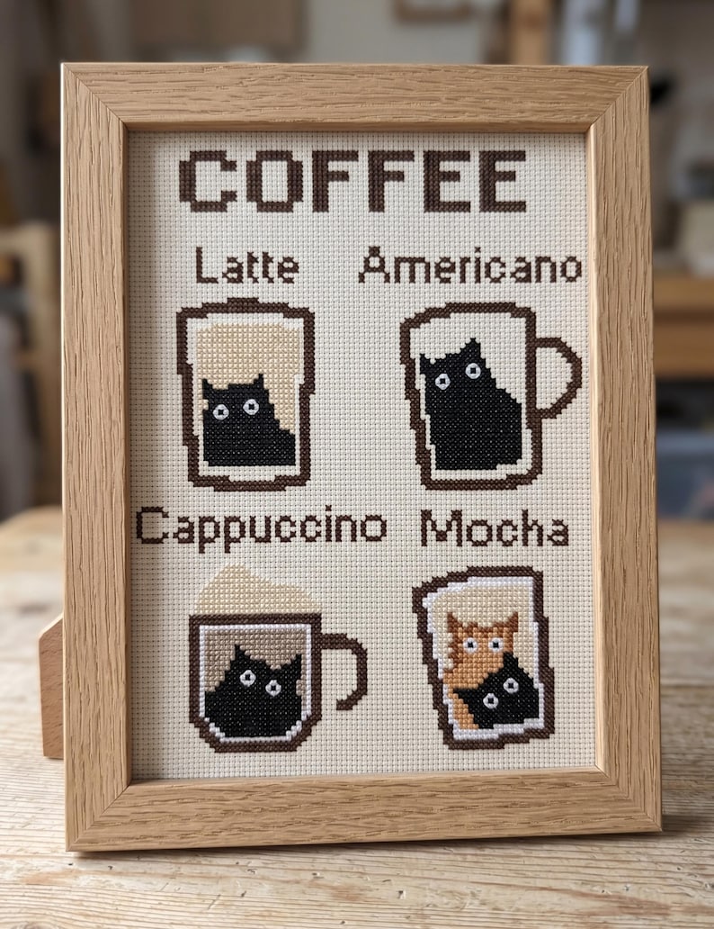 May include: Cross-stitch artwork in a light wood frame. The artwork features the word "COFFEE" above four coffee drink illustrations: Latte, Americano, Cappuccino, and Mocha. Each drink contains a cat.
