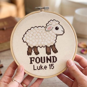K&ouml;nnte beinhalten: Ein runder Stickrahmen aus Holz mit einem Kreuzstichmotiv eines wei&szlig;en Schafs mit braunen Beinen und einem rosa Ohr. Unter dem Schaf steht der Text "FOUND" und "Luke 15" in dunkelbraunen Kreuzstichbuchstaben auf einem cremefarbenen Stoff.