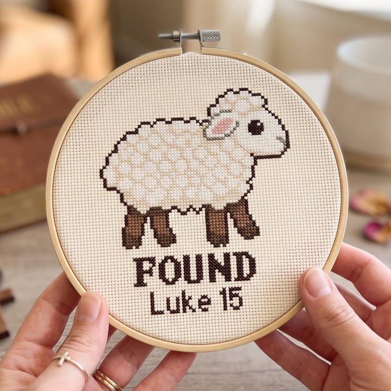 K&ouml;nnte beinhalten: Ein runder Stickrahmen aus Holz mit einem Kreuzstichmotiv eines wei&szlig;en Schafs mit braunen Beinen und einem rosa Ohr. Unter dem Schaf steht der Text "FOUND" und "Luke 15" in dunkelbraunen Kreuzstichbuchstaben auf einem cremefarbenen Stoff.