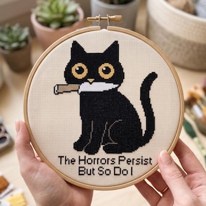 May include: A circular wooden embroidery hoop containing a cross-stitch design of a black cat with large yellow eyes holding a white knife in its mouth. Below the cat, the text reads: The Horrors Persist But So Do I in black lettering.