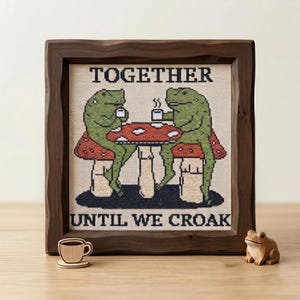 Könnte beinhalten: Gerahmtes Kreuzstich-Kunstwerk in dunklem Holz. Es zeigt zwei grüne Frösche, die an einem Pilztisch Kaffee trinken. Der Text lautet "TOGETHER UNTIL WE CROAK". Eine Holz-Kaffeetasse und eine Froschfigur sind ebenfalls vorhanden.