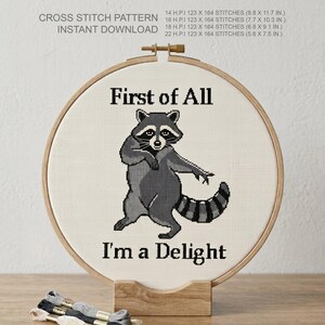 May include: A cross-stitch pattern featuring a raccoon with the text "First of All I'm a Delight." The design is displayed in a wooden embroidery hoop. The pattern includes stitch counts and dimensions in cm.