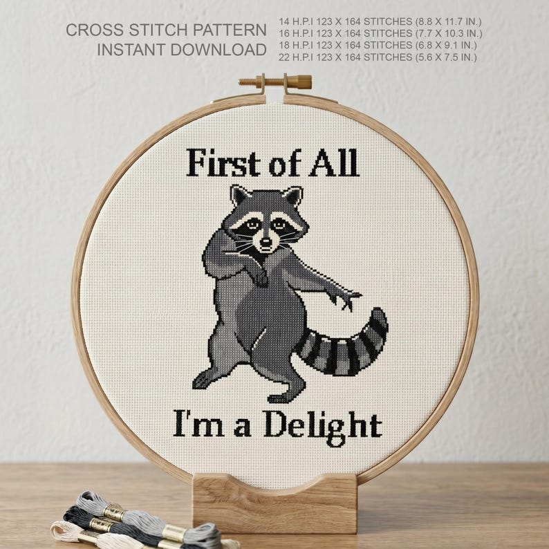 May include: A cross-stitch pattern featuring a raccoon with the text "First of All I'm a Delight." The design is displayed in a wooden embroidery hoop. The pattern includes stitch counts and dimensions in cm.
