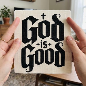 Peut inclure: Un carr&eacute; de tissu Aida blanc cass&eacute; pr&eacute;sentant un motif en point de croix noir. Le texte "God is Good" est brod&eacute; dans une police gothique grasse, avec une petite croix centr&eacute;e au-dessus du mot "God" et des &eacute;toiles d&eacute;coratives autour de "is".