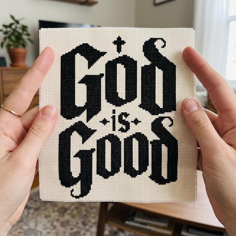 Peut inclure: Un carr&eacute; de tissu Aida blanc cass&eacute; pr&eacute;sentant un motif en point de croix noir. Le texte "God is Good" est brod&eacute; dans une police gothique grasse, avec une petite croix centr&eacute;e au-dessus du mot "God" et des &eacute;toiles d&eacute;coratives autour de "is".