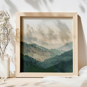 May include: A framed cross-stitch landscape depicting a mountain range in green, blue, and beige hues. The artwork is presented in a light-coloured wooden frame. The image suggests a natural, tranquil scene.