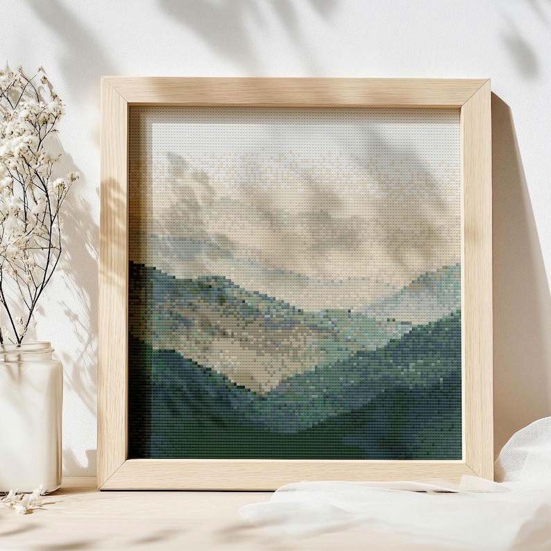 May include: A framed cross-stitch landscape depicting a mountain range in green, blue, and beige hues. The artwork is presented in a light-coloured wooden frame. The image suggests a natural, tranquil scene.