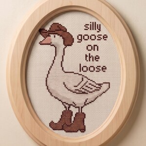 Op de afbeelding: Ovaalvormig kruissteekkunstwerk in een houten lijst. Het toont een gans met een cowboyhoed en laarzen. De tekst luidt "silly goose on the loose". De lijst is van licht hout met een leren ophanglus.