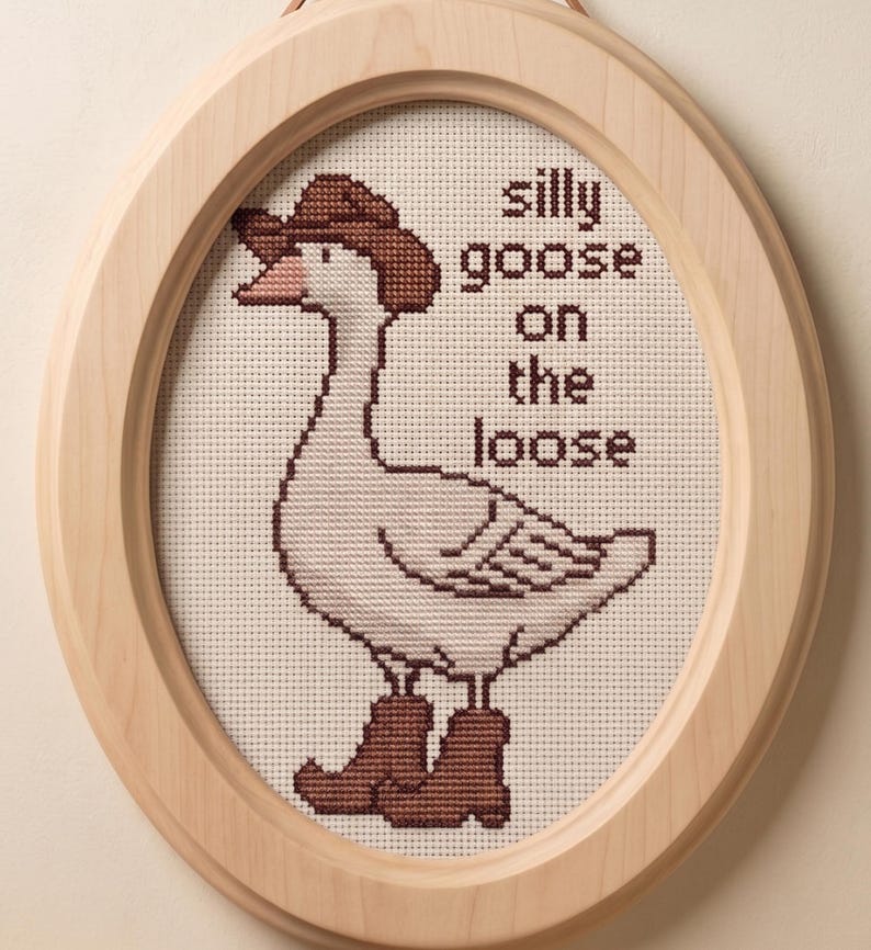 Op de afbeelding: Ovaalvormig kruissteekkunstwerk in een houten lijst. Het toont een gans met een cowboyhoed en laarzen. De tekst luidt "silly goose on the loose". De lijst is van licht hout met een leren ophanglus.