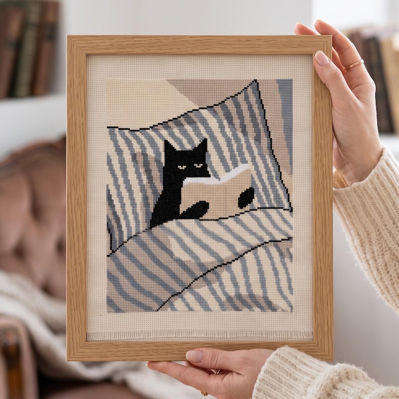 Op de afbeelding: Een ingelijst kruissteek borduurwerk met een zwarte kat die in bed onder een blauw-wit gestreepte deken ligt en een open boek leest. Het kunstwerk is geplaatst in een lichtgekleurde houten lijst en toont een gedetailleerd handwerkontwerp.