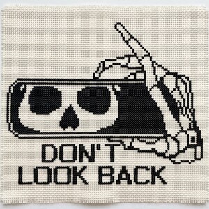 May include: A square cross-stitch embroidery on off-white fabric featuring a black skeleton hand holding a rectangular mirror with a skull reflection. The text DON'T LOOK BACK is stitched in black block letters below the mirror design.