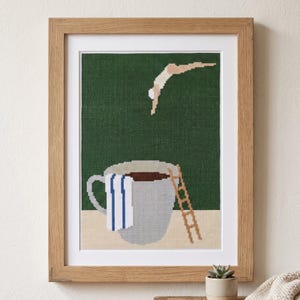 Op de afbeelding: Een ingelijst kruissteek kunstwerk met een donkergroene achtergrond en een figuur die in een grote grijze koffiemok duikt. De mok heeft een witte handdoek met blauwe strepen aan de zijkant en een bruine ladder die op een beige ondergrond staat.