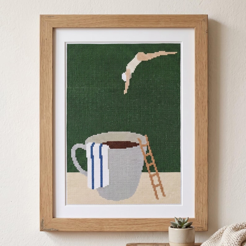 Puede incluir: Una obra de punto de cruz enmarcada con un fondo verde oscuro que muestra una figura saltando a una gran taza de caf&eacute; gris. La taza tiene una toalla blanca con rayas azules y una escalera marr&oacute;n apoyada sobre una superficie de color beige.