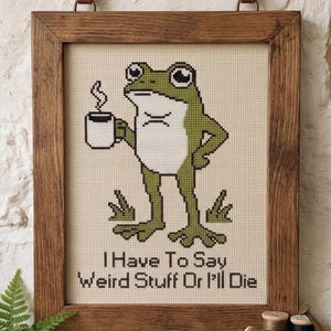 Puede incluir: Un patr&oacute;n de punto de cruz en un marco de madera que muestra una rana verde sosteniendo una taza blanca humeante. El texto debajo de la rana dice, I Have To Say Weird Stuff Or I'll Die. El dise&ntilde;o est&aacute; sobre una tela color crema con cuadr&iacute;cula.