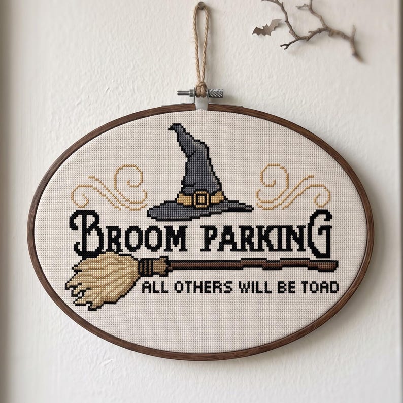 May include: An oval cross-stitch embroidery in a wooden hoop featuring a grey witch hat, a tan broom, and the black text "Broom Parking" and "All others will be toad" on a cream fabric background, suspended by a jute twine loop.