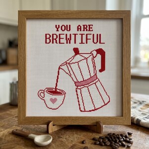 K&ouml;nnte beinhalten: Ein quadratisches Kreuzstich-Stickbild in einem hellen Holzrahmen zeigt eine rote Mokkakanne, die Kaffee in eine Tasse mit einem kleinen rosa Herz gie&szlig;t. Der Text dar&uuml;ber lautet YOU ARE BREWTIFUL in roten Buchstaben auf wei&szlig;em Stoff.