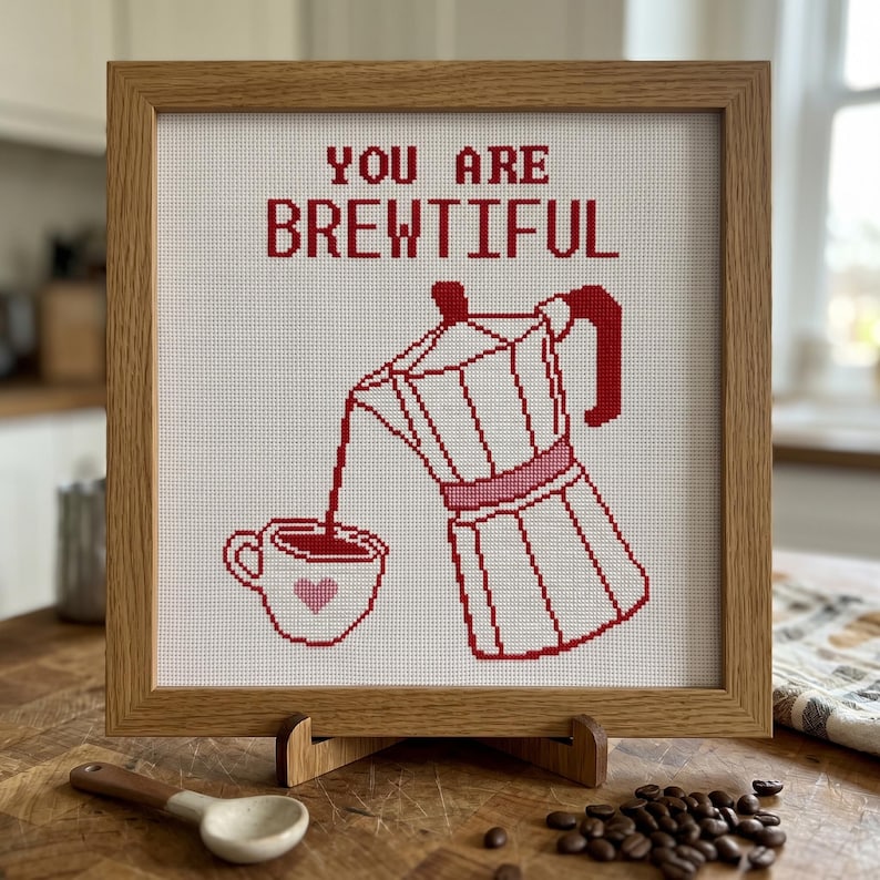 K&ouml;nnte beinhalten: Ein quadratisches Kreuzstich-Stickbild in einem hellen Holzrahmen zeigt eine rote Mokkakanne, die Kaffee in eine Tasse mit einem kleinen rosa Herz gie&szlig;t. Der Text dar&uuml;ber lautet YOU ARE BREWTIFUL in roten Buchstaben auf wei&szlig;em Stoff.