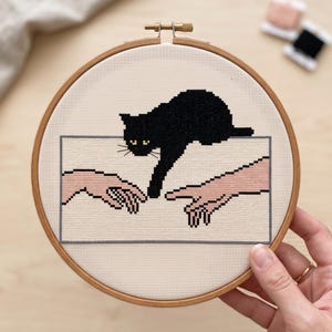 Pode incluir: Um bordado em ponto cruz num bastidor de madeira circular apresentando um gato preto estendendo uma pata em dire&ccedil;&atilde;o a uma m&atilde;o humana. O desenho est&aacute; sobre um tecido creme claro e emoldurado por uma fina borda retangular cinza.