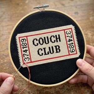 May include: A round wooden embroidery hoop containing black fabric with a cross-stitched design of a cream-coloured ticket with red borders. The ticket features the black text COUCH CLUB and the number 374189 on both ends. A needle and thread are visible.