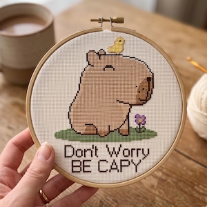 May include: A circular wooden embroidery hoop containing a cross-stitch design of a light brown capybara with a small yellow bird on its head, a purple flower on green grass, and the text "Don't Worry BE CAPY" stitched in dark brown thread.