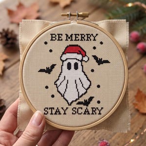 K&ouml;nnte beinhalten: Eine Kreuzstichstickerei in einem runden Holzrahmen zeigt ein wei&szlig;es Gespenst mit roter Weihnachtsmannm&uuml;tze, umgeben von schwarzen Flederm&auml;usen und Punkten. Der Text "BE MERRY" ist in Schwarz dar&uuml;ber und "STAY SCARY" in Schwarz darunter gestickt.