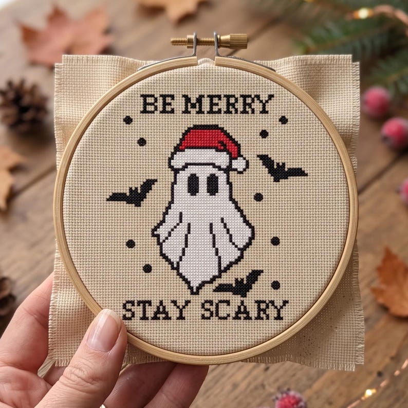 K&ouml;nnte beinhalten: Eine Kreuzstichstickerei in einem runden Holzrahmen zeigt ein wei&szlig;es Gespenst mit roter Weihnachtsmannm&uuml;tze, umgeben von schwarzen Flederm&auml;usen und Punkten. Der Text "BE MERRY" ist in Schwarz dar&uuml;ber und "STAY SCARY" in Schwarz darunter gestickt.